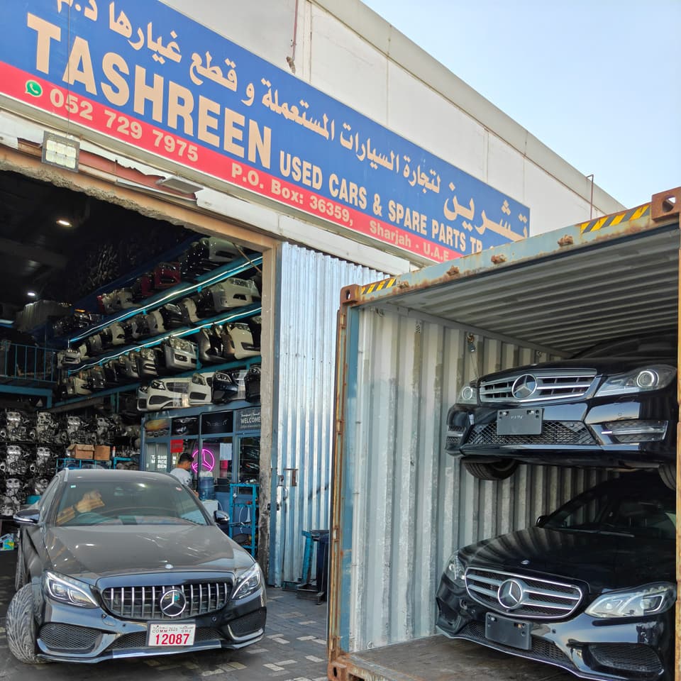 واجهة محل تشرين في الشارقة تحت لافتة الشركة، وفي الداخل رفوف واجهات أمامية، وأمامه سيارة مرسيدس-بنز سوداء واثنتان أخريان داخل حاوية شحن